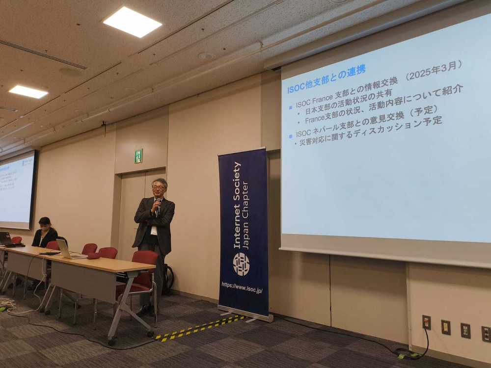 A man stands next to a projector screen and speaks into a microphone at a Japan Chapter event